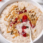 brown rice porridge in a white bowl drizzled with peanut butter and sprinkled with raspberries. The bowl is on a white backdrop.