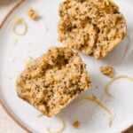 lemon poppyseed muffin cut in half showcasing the soft interior. The muffin is on a white plate and sprinkled with lemon zest.