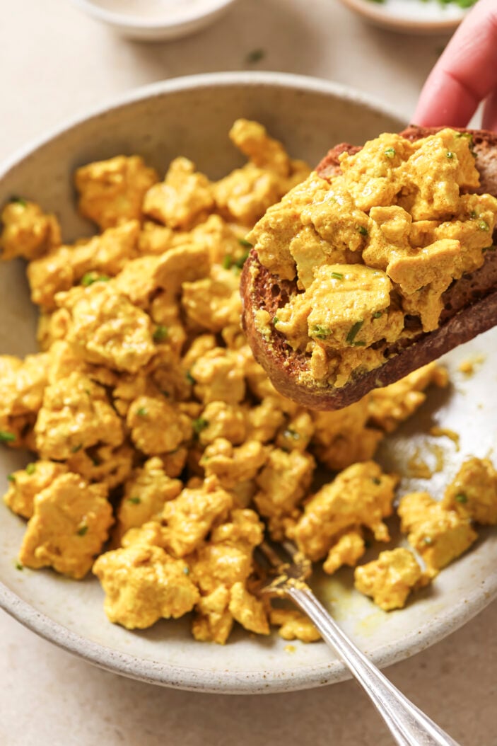 tofu scrambled scooped onto toast with the white bowl of scramble in the background.