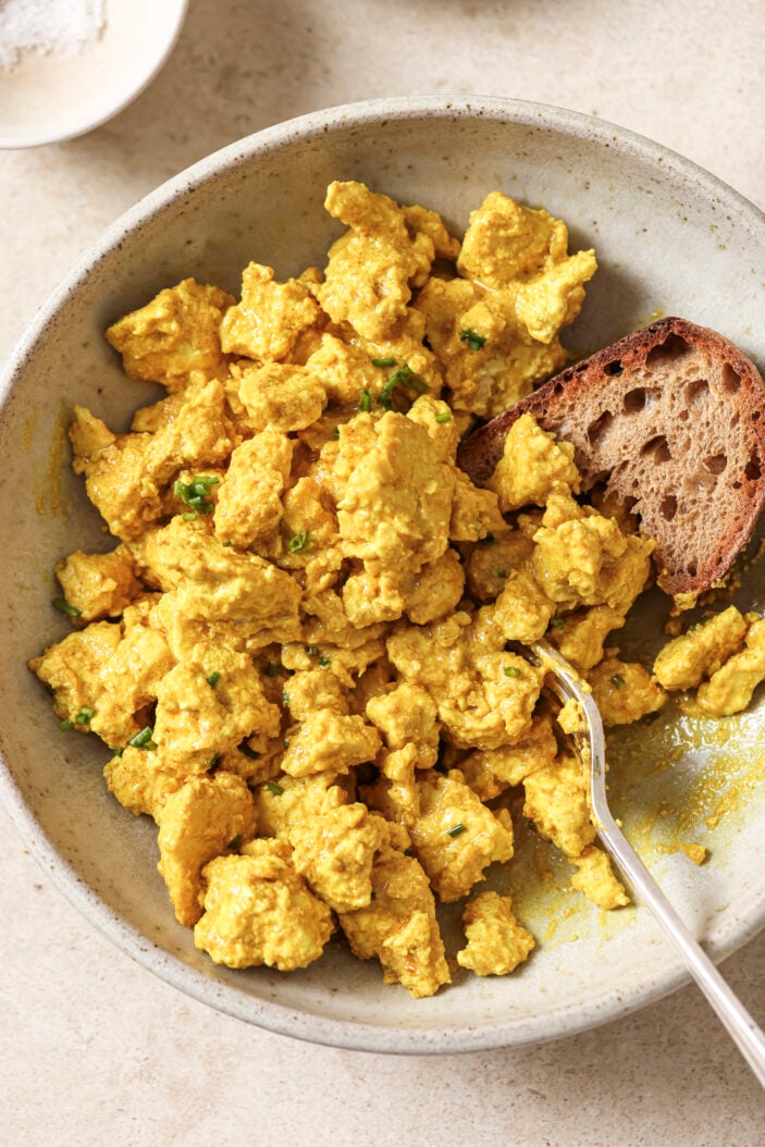 top down image of vegan tofu scramble in a white bowl with a side of toast and a metal spoon in th ebowl. Showcasing eggy and creamy consistency.