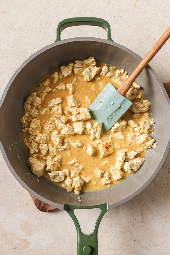 tofu crumbled into a skillet with oat milk and spices mixed together and ready to simmer. A wooden spatula is in the middle.