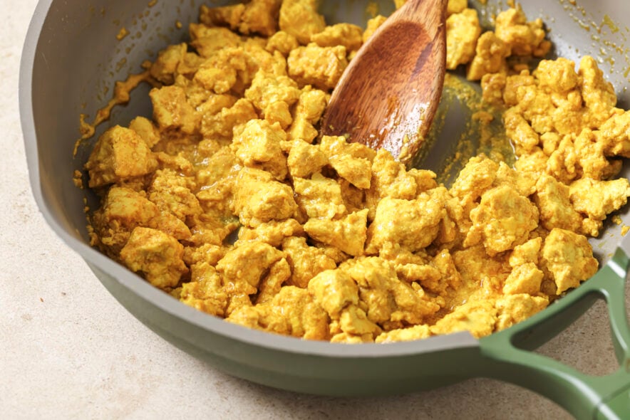Side shot of bright yellow tofu scramble showcasing eggy consistency in a skillet with a wooden spoon in the center of the skillet.