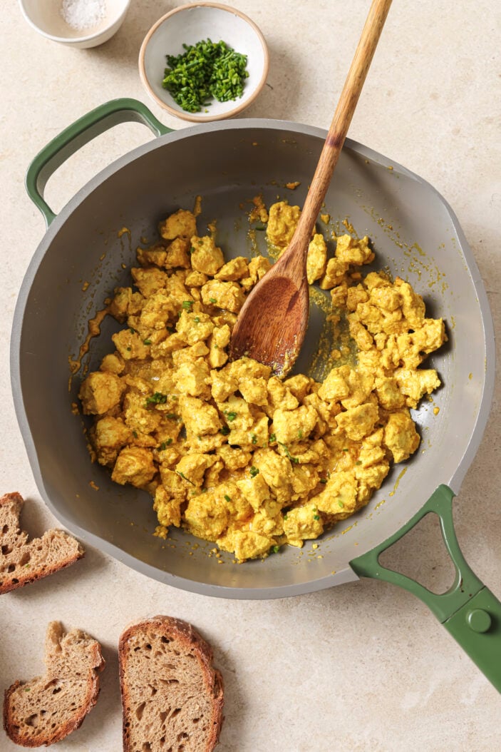 Tofu scramble that's bright yellow and ready to eat in a skillet with a wooden spoon the middle and pieces of toasted bread on the side.