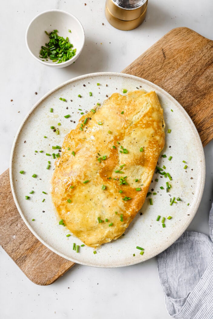 tofu omlette on a white plate stuffed with vegan cheese, spinach and mushroom. Sprinkled with fresh chopped chives. The omelette is on a wood cutting board and there is a small dish of chives in the top left corner.