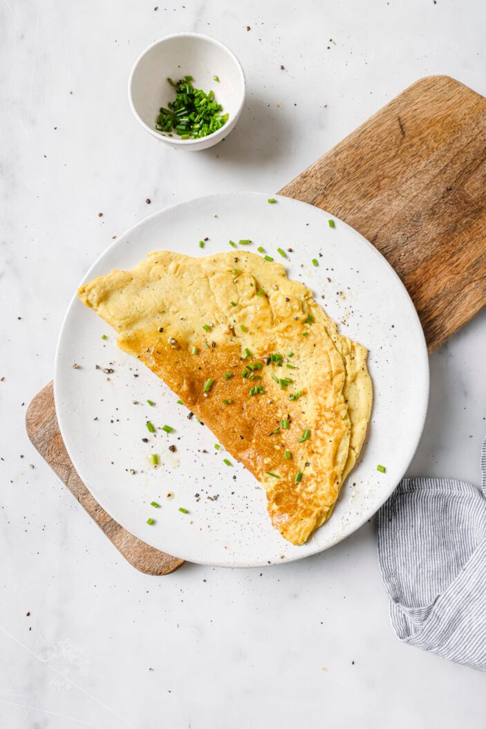 tofu omelette served on a white plate on top of a wood cutting board. There is a small bowl of chopped chives in the top left corner.