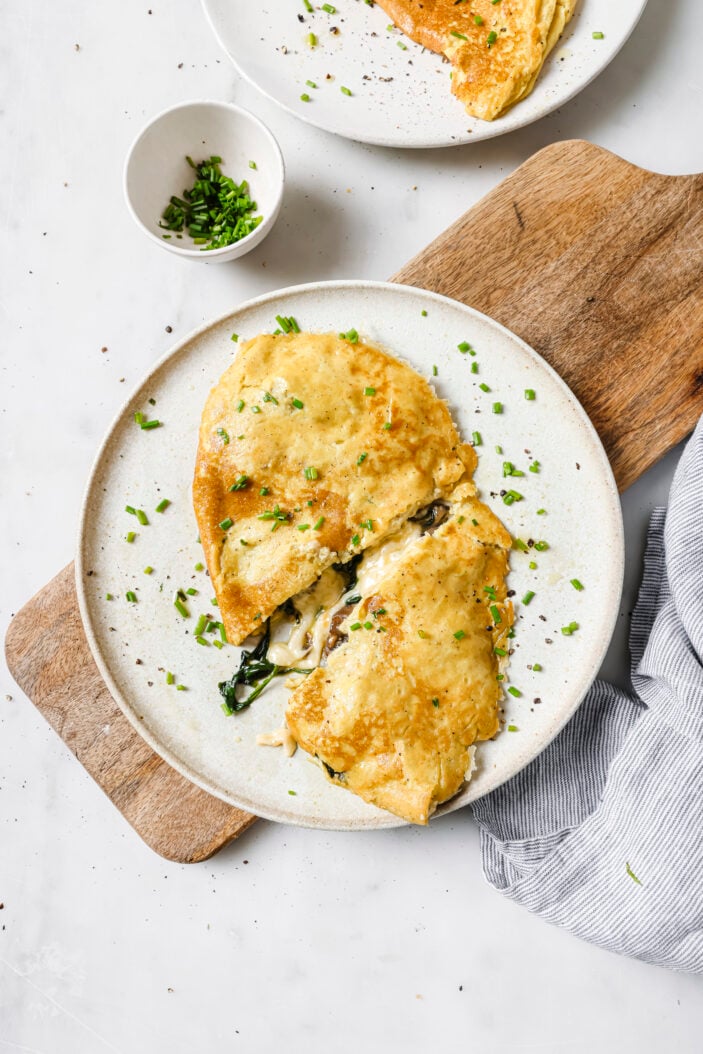 tofu omlette on a white plate stuffed with vegan cheese, spinach and mushroom. The omelette is sliced in half and sprinkled with fresh chopped chives. The omelette is on a wood cutting board and there is a small dish of chives in the top left corner.