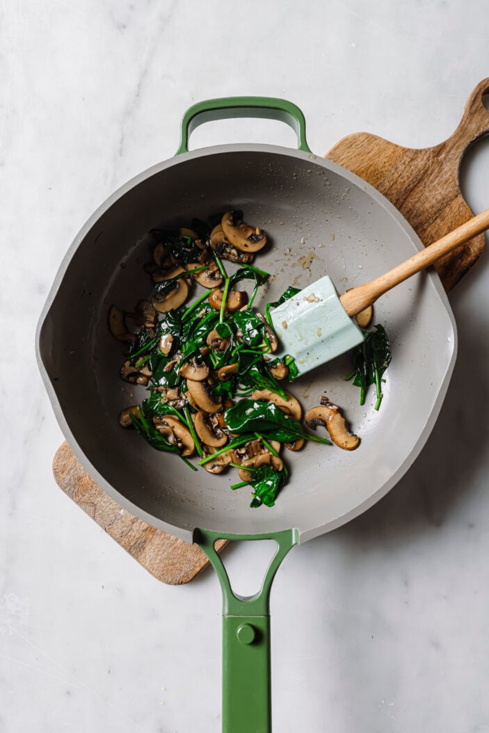 Cooked mushrooms and wilted spinach in a skillet with a spatula in the middle.