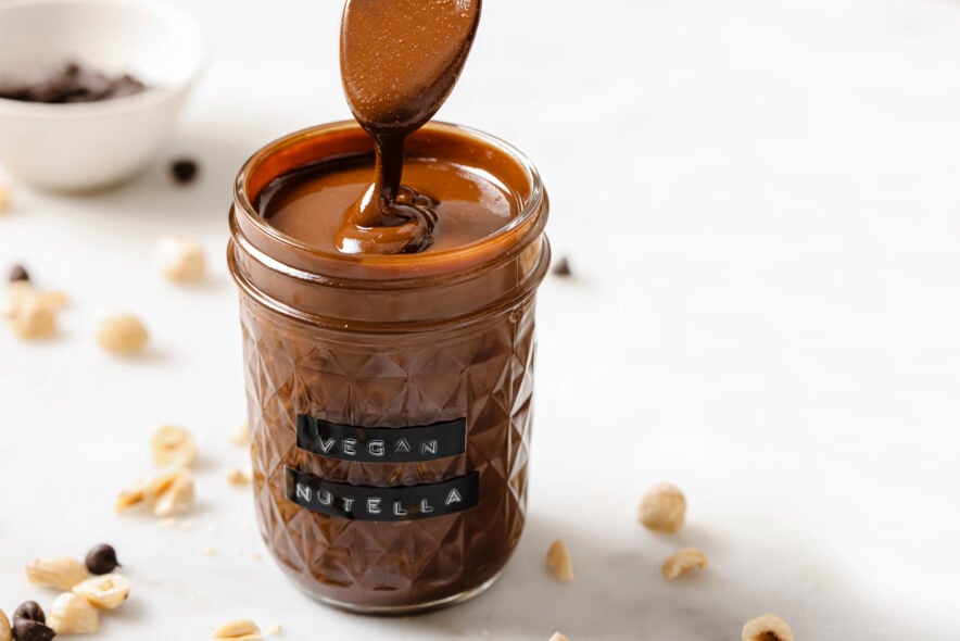 vegan nutella in a clear glass jar for storage. A spoon is being scooped into nutella showcasing its smooth and drizzly texture.