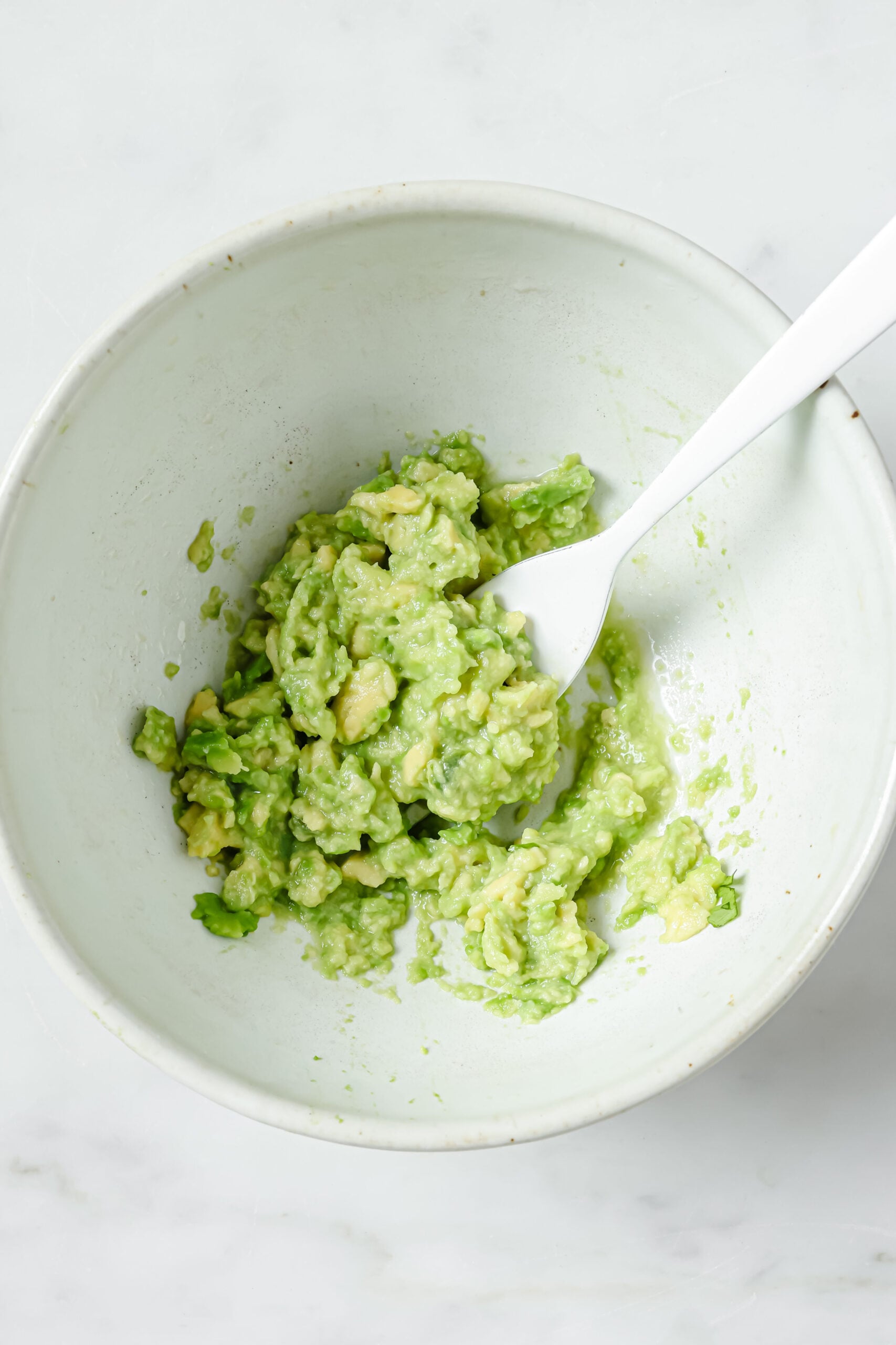 A simple guacamole in a white bowl that's been made by mashing avocado with a fork and adding salt and lime juice. The white bowl is sitting on a white marble backdrop.