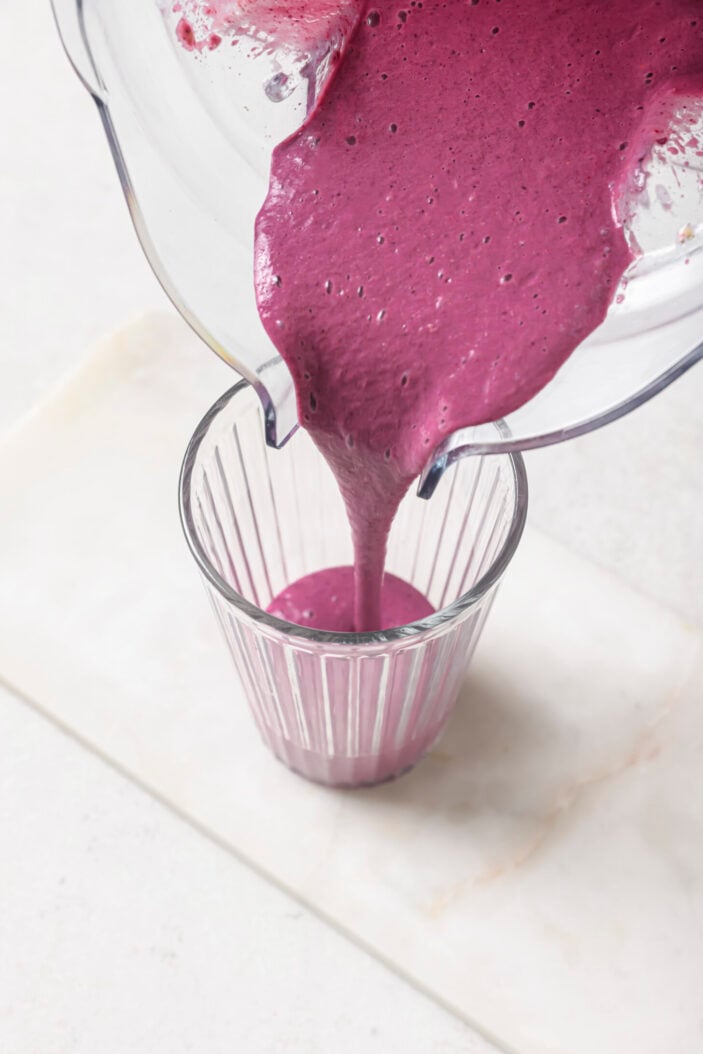 Pouring berry banana smoothie from a blender container into a large tall glass. This showcases the smooth and creamy consistency.