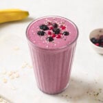 image of berry banana smoothie in a tall glass, decorated with frozen berries on top and a sprinkling of chia seeds and hemp hearts