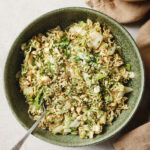 image of nappa cabbage salad with ramen noodles in a green bown with a fork in it. Sprinkled with fresh green onion and cilantro. There is a brown kitchen towel to the left side of the bowl.