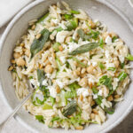 A ceramic bowl filled with lemon orzo pasta salad, chickpeas, crumbled feta cheese, green herbs, and toasted pine nuts, with a silver spoon resting inside. The dish sits on a light-colored surface.