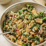 side angle image of a warm kale and crispy tofu salad in a large bowl with a silver fork in the middle. In the back corner is a small bowl of cashews and a wood cutting board.