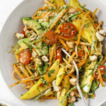 A bowl of colorful salad with sliced mango, avocado, cherry tomatoes, spiralized carrots, cashews, and herbs, with a fork resting inside. A small bowl of chopped herbs is visible above the main bowl.