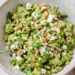 A bowl of green quinoa salad with avocado, cashews, crumbled feta cheese, cucumber, and fresh herbs, served in a light-colored bowl.