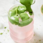 A glass iced matcha coconut cloud - with a layer of pink iced coconut water is being topped with a thick, green matcha cold foam poured from another glass.