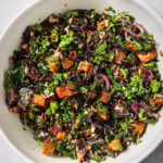 A white bowl filled with a colorful salad of chopped kale, roasted sweet potatoes, wild rice, red onion, dried cranberries, and nuts, all mixed together on a light gray background.
