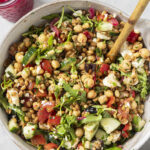 A bowl of mixed salad with chickpeas, cucumbers, tomatoes, greens, feta, and grains, with a wooden spoon. A jar with pickled onions is nearby on a light surface.
